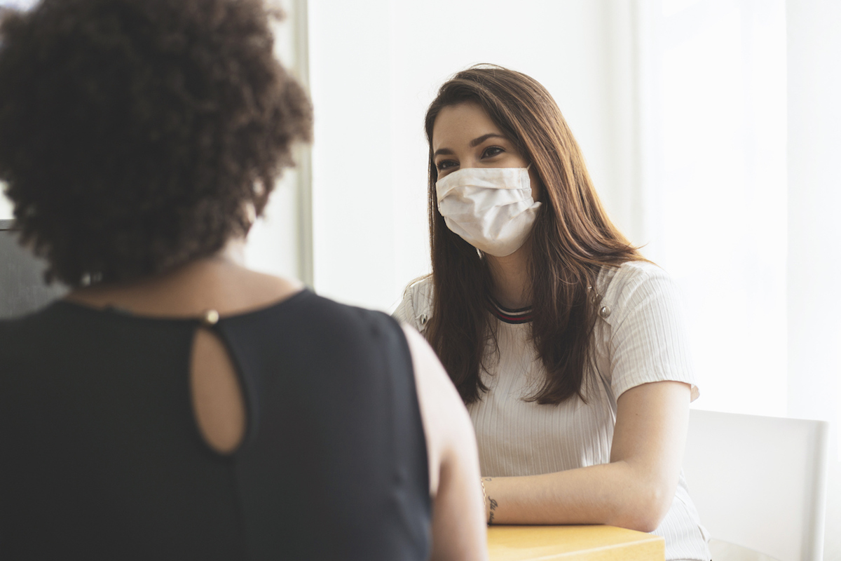 Two Young Women Talking In An Office Wearing A Protective Face Mask.