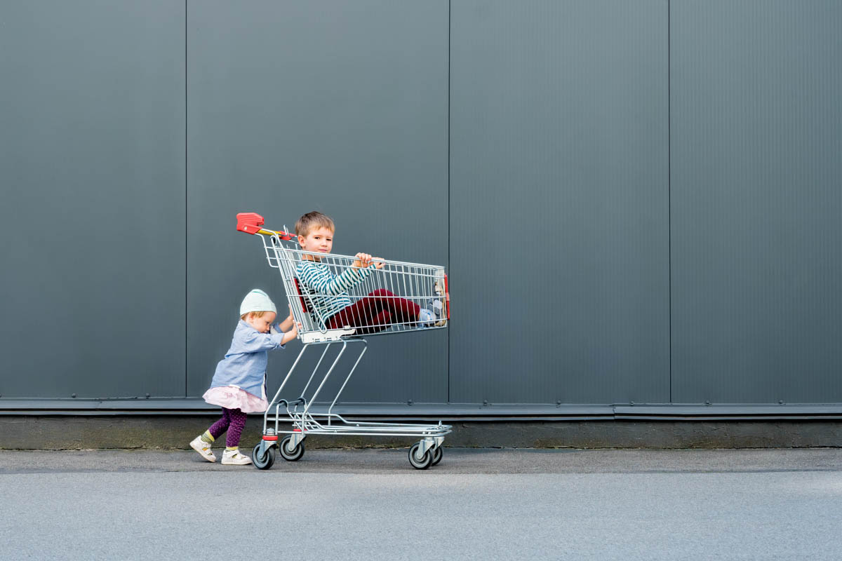 two trendy hipsters with shopping cart near supermarket, shopping mall. two cute children waiting for parents outdoor. happy childhood concept background. little girl playing with her brother