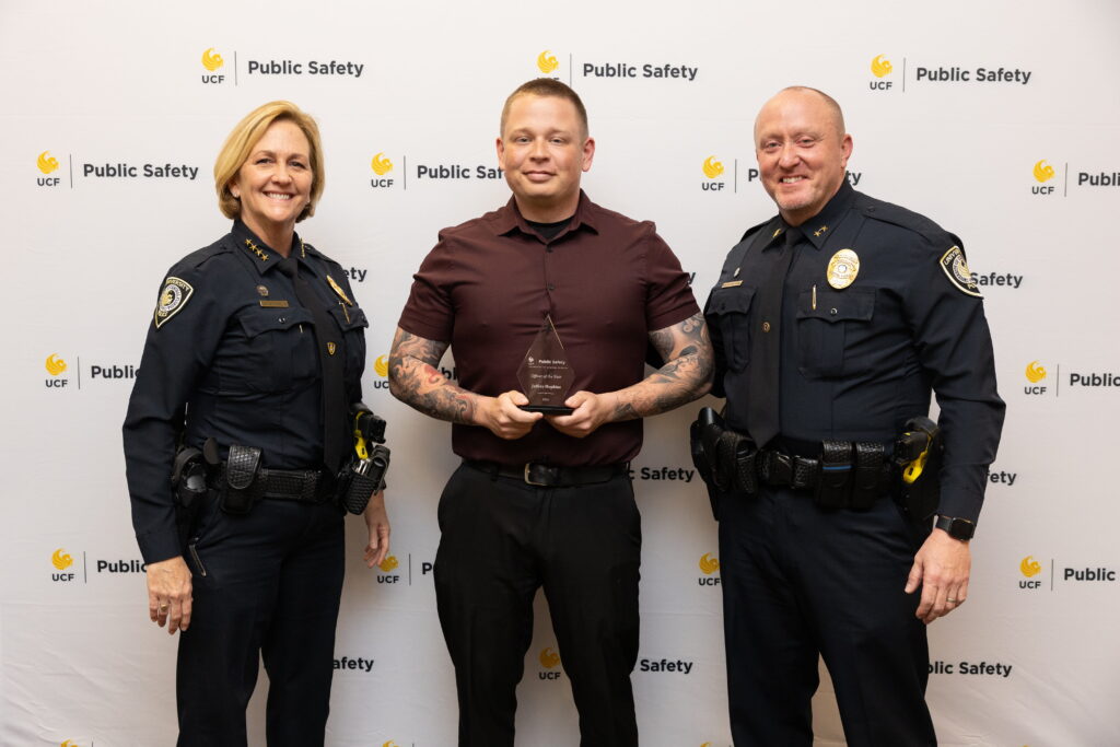 Corporal Jeffrey Hopkins standing with Associate Vice President of Public Safety and Chief of Police Robin Griffin-Kitzerow and Deputy Chief of Police Scott Freeman  while holding his award at the UCF Public Safety Awards ceremony. 