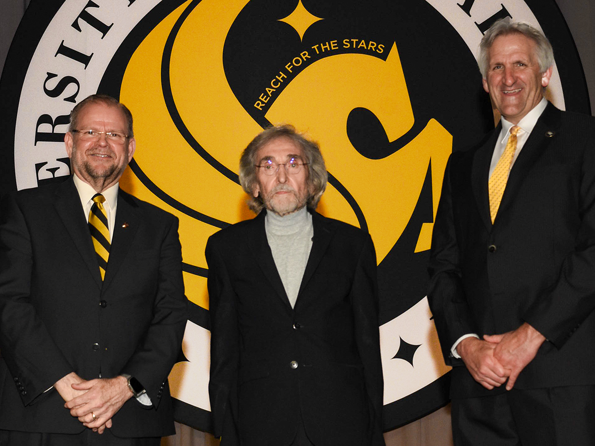 UCF President (left) and Provost (right) stand on stage with faculty honoree Charles “Chuck” Dziuban at center during the Founders’ Day Faculty Celebration, with the UCF seal displayed in the background.