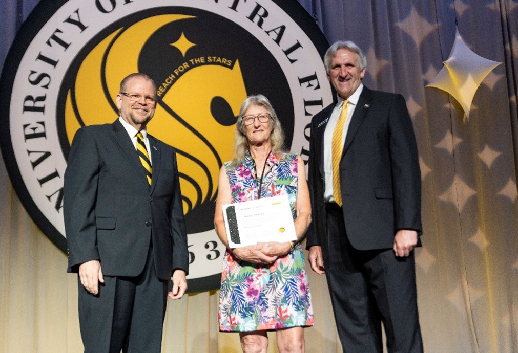 UCF President (left) and Provost (right) stand on stage beside the employee with the most years of service, Donna Hickson (center), who holds her 45 years of service certificate, with the UCF seal prominently displayed in the background.