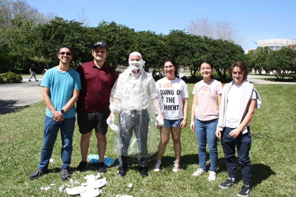 Outdoor group photo featuring Dr. Jeff Kauffman covered in whipped cream during a “Pie a Prof” campus event while standing with students on a grassy lawn at the University of Central Florida.