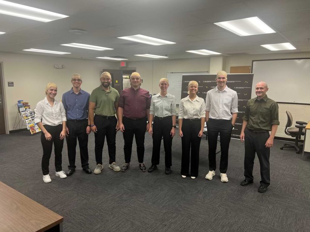 Indoor group photo featuring students dressed to resemble Dr. Jeff Kauffmann during a Halloween event at the University of Central Florida. The students are standing in a classroom space wearing similar outfits and bald caps, with Dr. Kauffmann standing at the far right of the group.