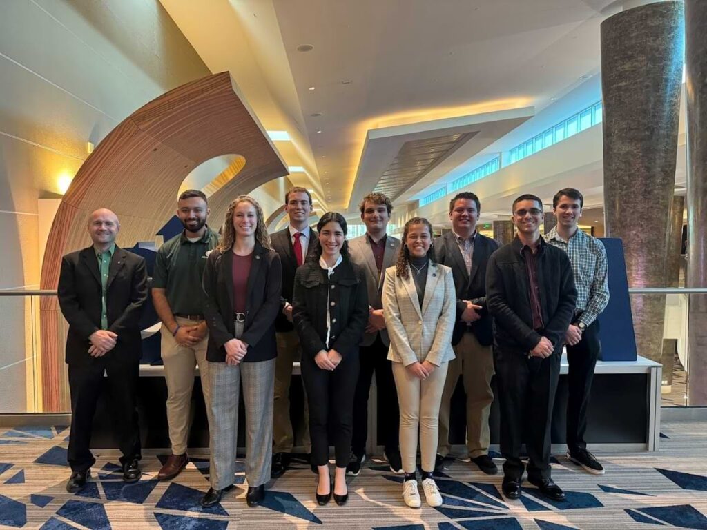 Group photo featuring Dr. Jeff Kauffmann (far left) standing with students at the 2025 AIAA SciTech Forum inside a large conference venue. The group is dressed in professional attire and posing together in front of a decorative display.