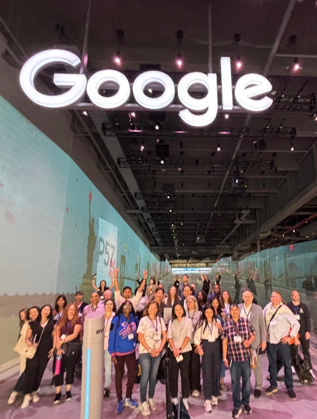 Group photo of faculty and participants standing beneath a large, illuminated Google sign during a visit to Google’s New York City offices.