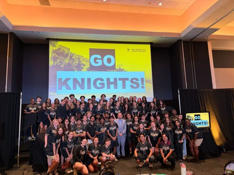 UCF President Alexander Cartwright poses with members of the New Student and Family Programs team in front of a large "Go Knights!" screen following the department's May 2026 training.