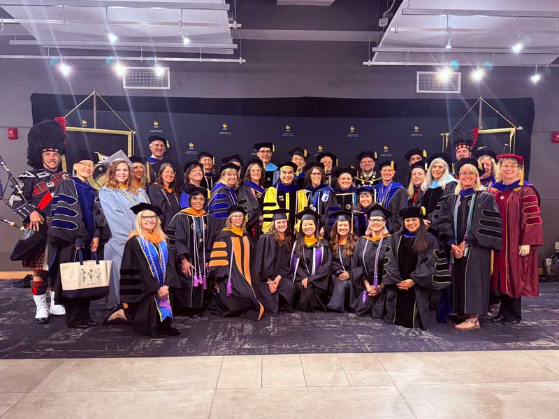 Dr. Michael Joseph Dino, wearing academic regalia, stands with a large group of College of Nursing faculty and colleagues dressed in academic regalia at graduation.