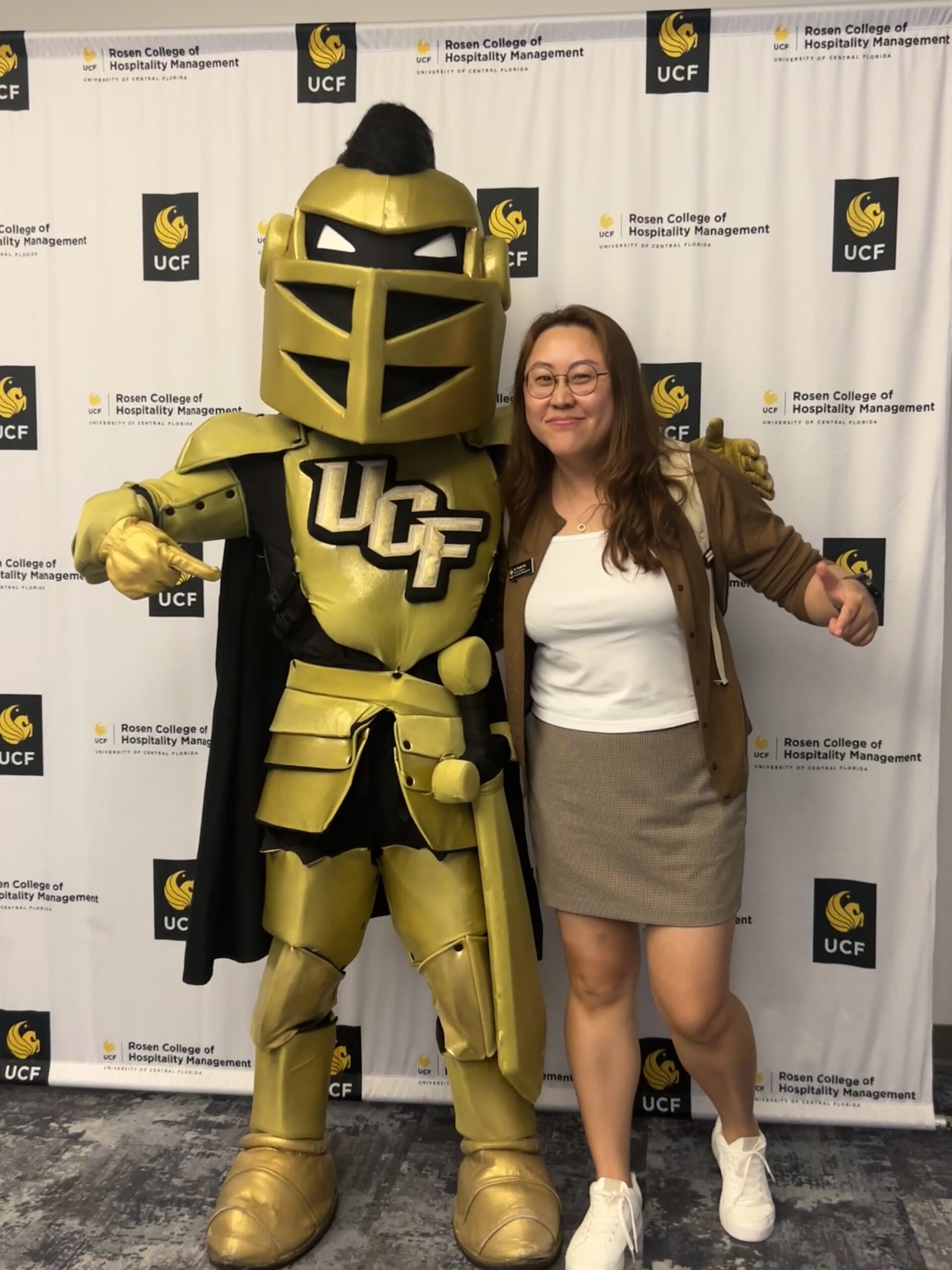 Dr. Forest Ma smiles and poses with Knightro in front of a Rosen College of Hospitality Management backdrop.