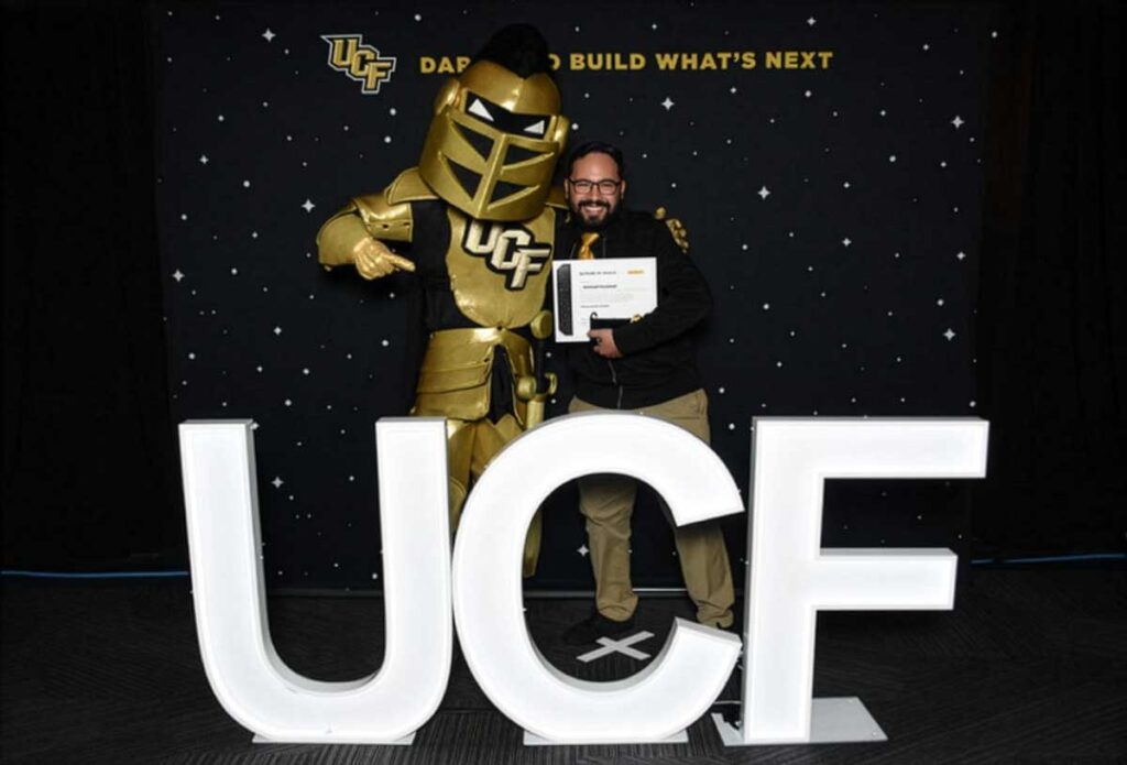 Knightro stands with Samuel Ensenat, who holds his 20-year service award certificate behind large, illuminated UCF letters.